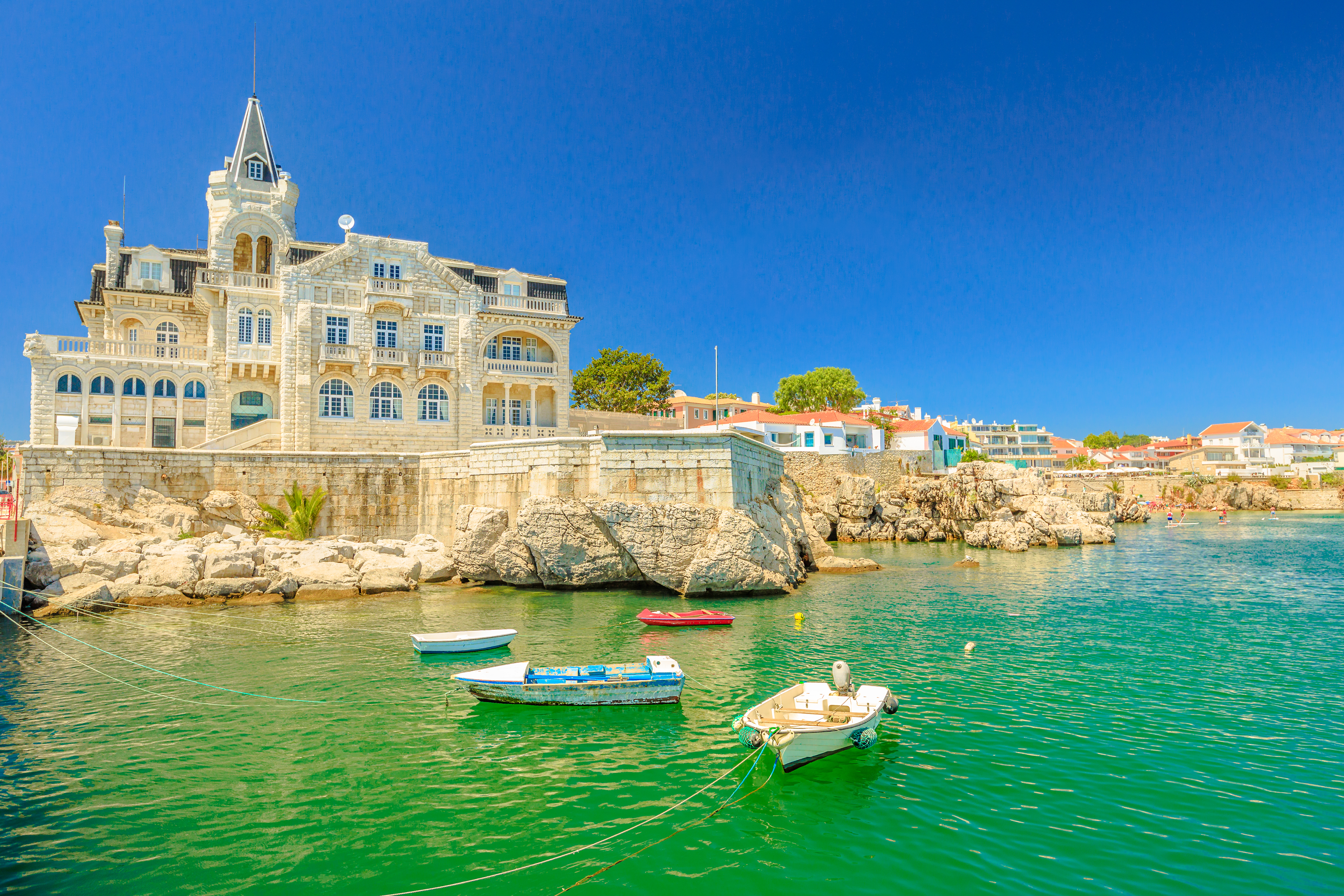 Idyllisk udsigt over det forladte Palace Seixas og farverige både ved strandpromenaden i Cascais, med turkisblåt hav og Praia da Rainha i baggrunden