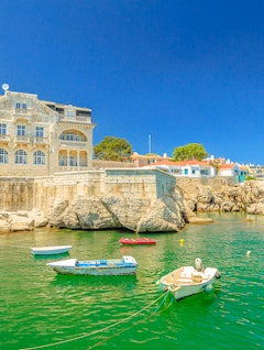 Idyllisk udsigt over det forladte Palace Seixas og farverige både ved strandpromenaden i Cascais, med turkisblåt hav og Praia da Rainha i baggrunden