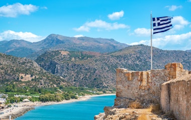 Historiske ruiner af Castel Selino med det græske flag der vajer over den idylliske kystby Paleochora med dens kridhvide huse, turkisblå bugt og bjerglandskab på Kreta.