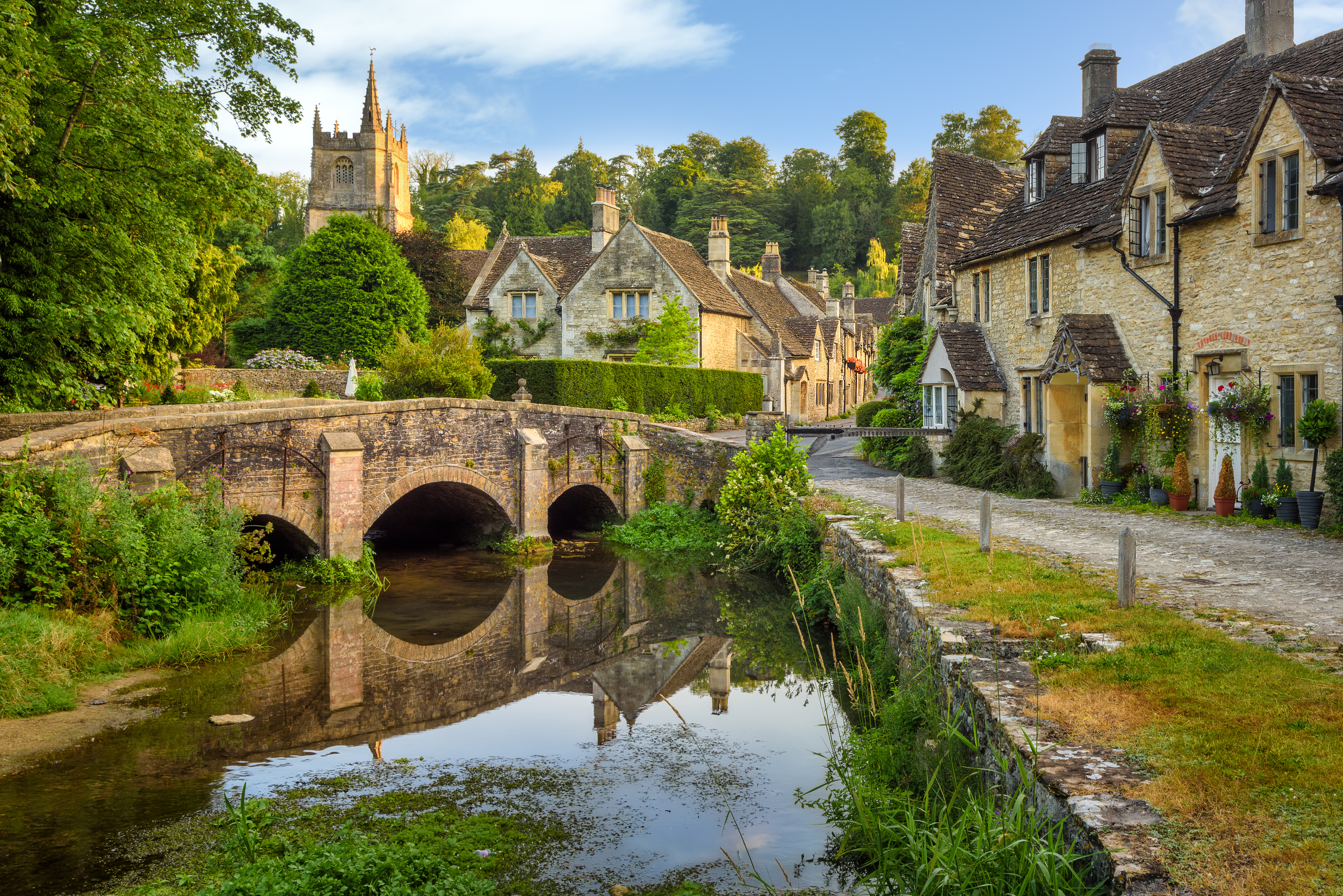 Idyllisk Castle Combe landsby i Cotswolds, England, med en middelalderlig stenbro der spejler sig i åen, traditionelle honningfarvede stenhuse langs den brolagte gade og et kirketårn der rejser sig over de grønne træer