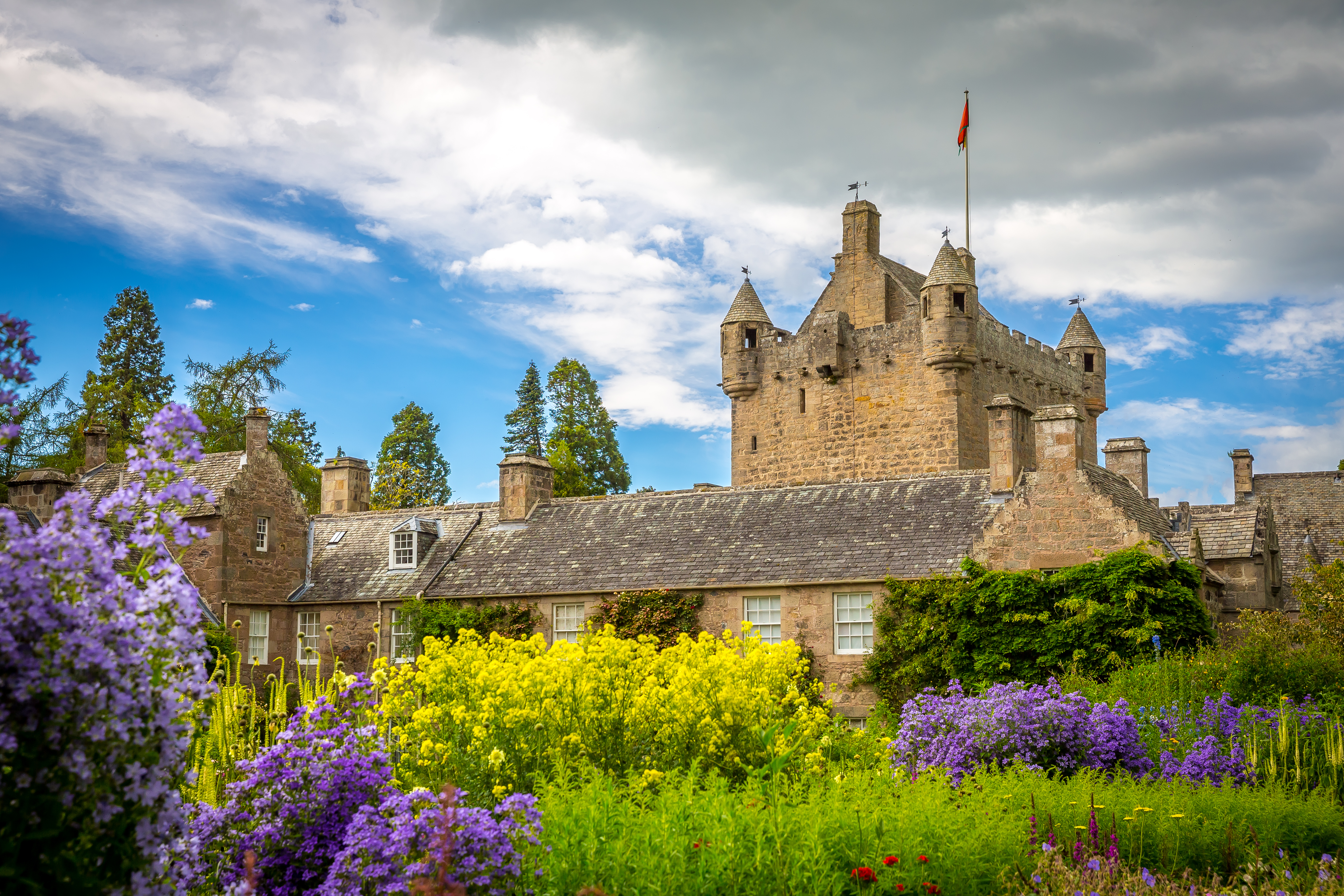 Cawdor Castle i de skotske højlande omgivet af farverige blomsterhaver med lilla og gule blomster under en dramatisk blå himmel