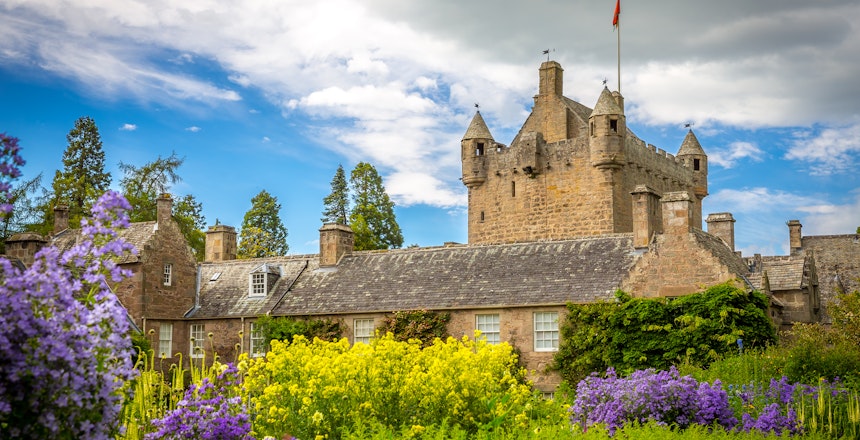 Cawdor Castle i de skotske højlande omgivet af farverige blomsterhaver med lilla og gule blomster under en dramatisk blå himmel
