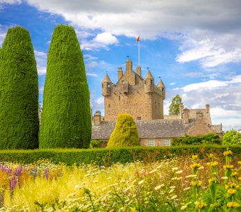 Cawdor Castle i de skotske højlande omgivet af smukke haver med vilde blomster og kegleformede topiary-træer under en blå sommerhimmel