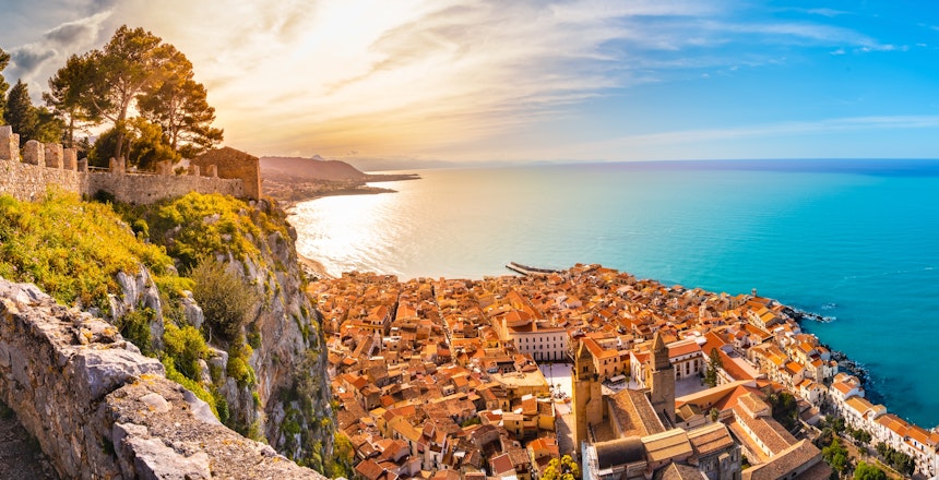 Panoramaudsigt over den historiske middelalderby Cefalù på Sicilien ved solnedgang med turkisblåt hav og bjerge i baggrunden
