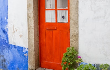 Smuk rød dør med hvide gardiner i Obidos, Portugal, omgivet af en stenbelagt veranda og en dekorativ blomstervase