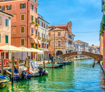 Malerisk udsigt over Vena-kanalen i Chioggia med farverige fortøjede både, historiske bygninger og en hyggelig restaurant ved vandkanten under klar blå himmel i Veneto, Italien