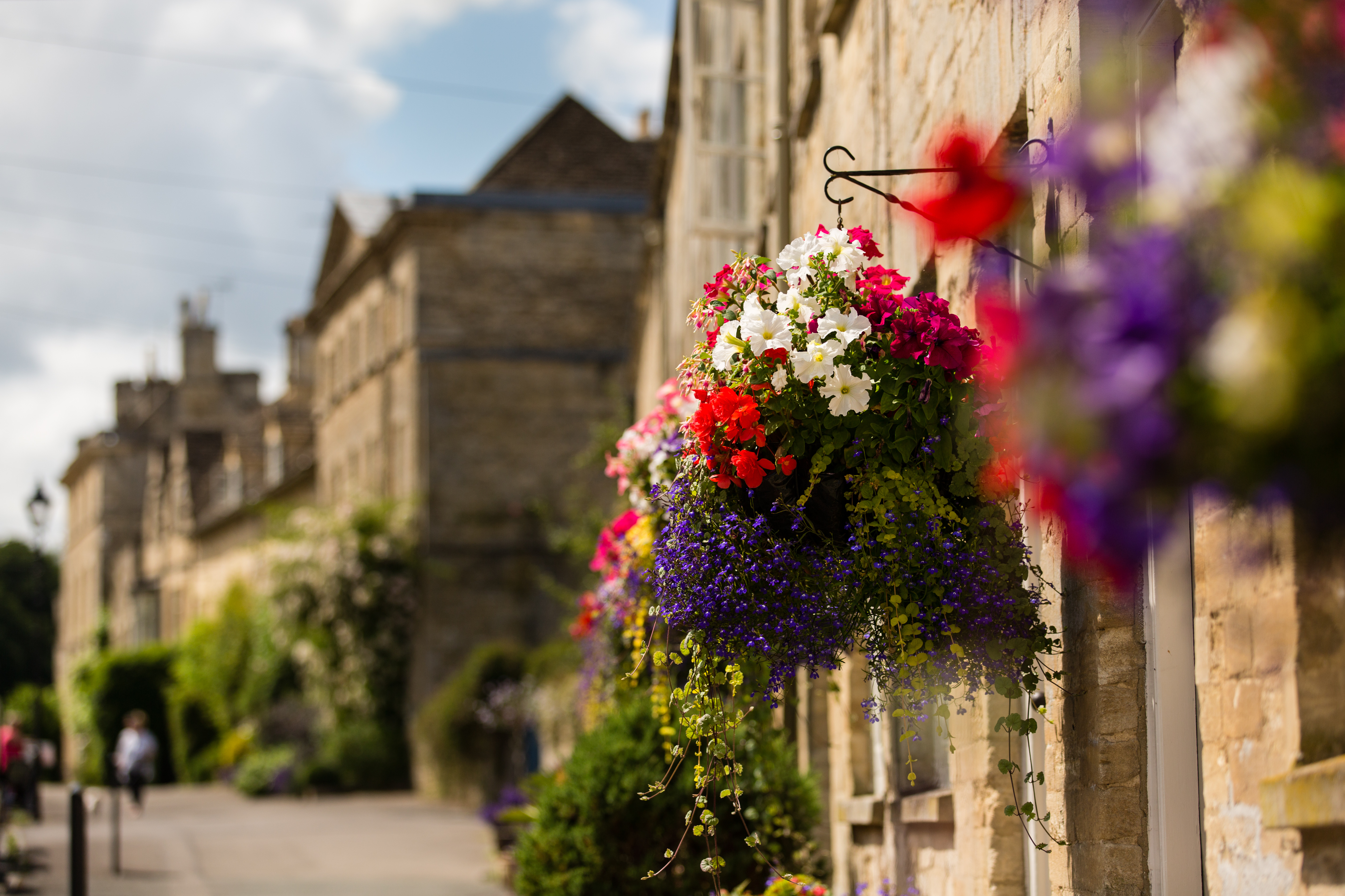 Hyggelig gade i Cirencester i Cotswolds med farverige hængende blomsterkurve på historiske kalkstensbyggerier
