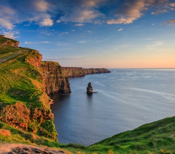 Betagende solnedgang ved Cliffs of Moher i Irland med dramatiske klipper, grønne skråninger og Atlanterhavets bølger mod den ikoniske klippesøjle