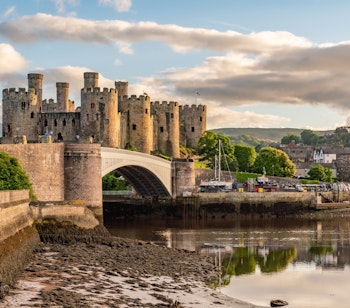Det imponerende middelalderslot Conwy i Nordwales, badet i gyldent aftenlys med solnedgangens orange og pink farver, der reflekteres i den stille flodmunding