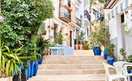Idyllisk gadebillede fra Costa Blanca med traditionelle hvidkalkede huse, blå detaljer, smedejerns-balkoner og farverige blomsterkrukker langs en charmerende trappe i Alicante
