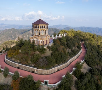Dronefoto af Throni of Panagia Kykkos klosteret på bjergtop i Cypern med hvide bygninger omgivet af grøn skov og bjerglandskab