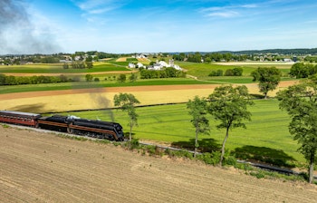 Betagende luftfoto af et historisk damptog der puster sort røg, mens det kører gennem et malerisk landskab med farverige marker, grønne bakker og landlig idyl på en nostalgisk togrejse