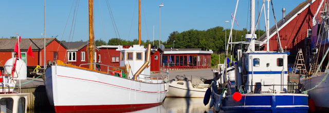 Traditionelle fiskerbåde i dansk havn med røde træhuse og blå himmel