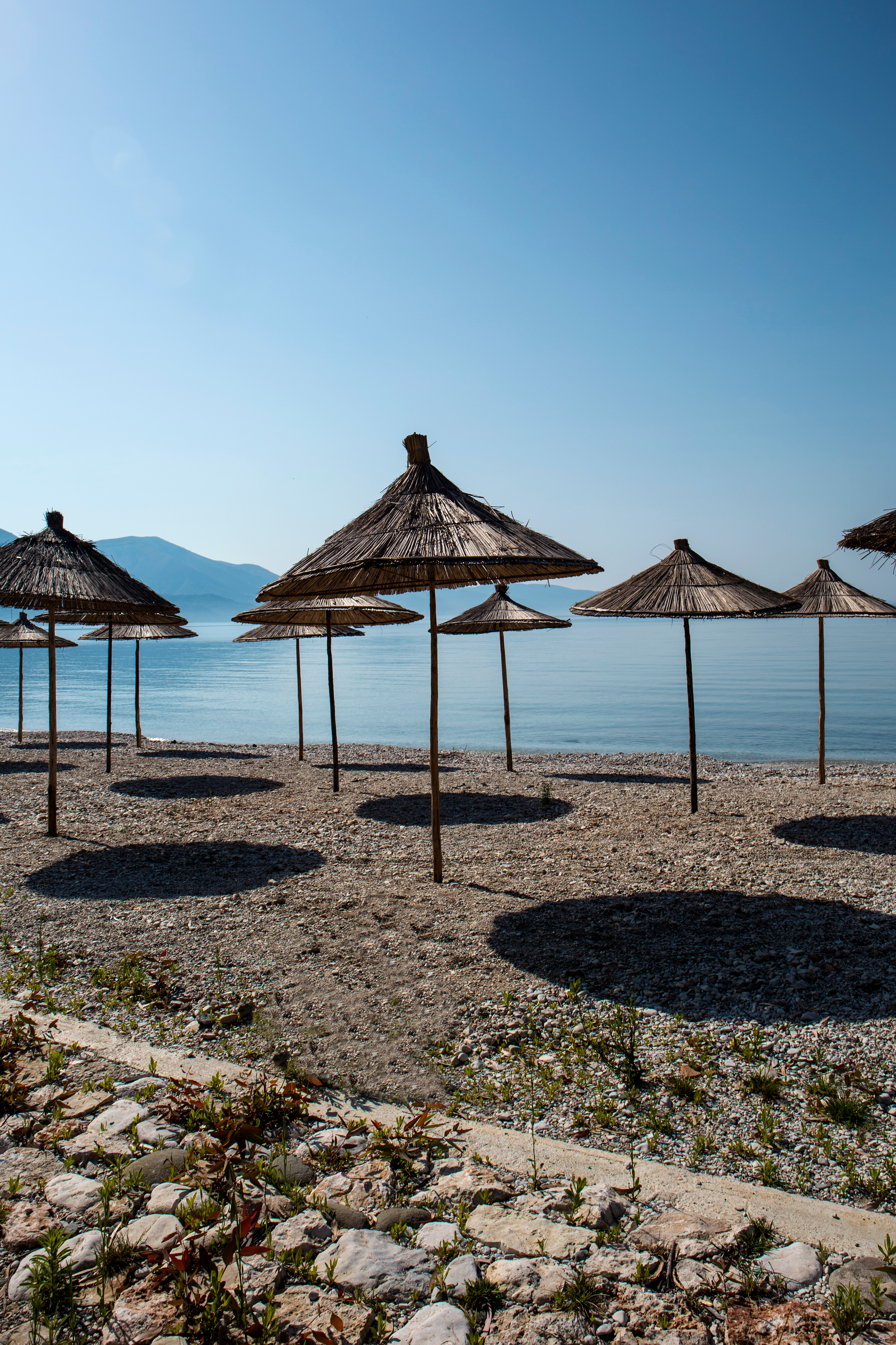 Farverige strandparasoller ved den idylliske kyst i Dhermi, Albanien, med krystalklart blåt vand og bjergkæder i baggrunden