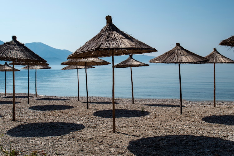 Farverige strandparasoller ved den idylliske kyst i Dhermi, Albanien, med krystalklart blåt vand og bjergkæder i baggrunden