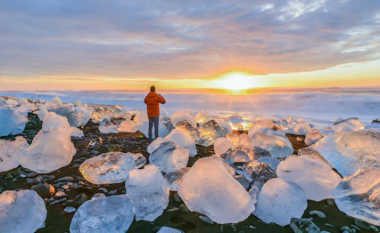 Person i orange jakke betragter solopgang ved Diamond Beach på Island med glitrende isblokke på den sorte vulkanske strand nær Jokulsarlon-gletsjerlagunen