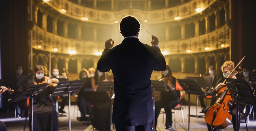 Dirigent leder symfoniorkester med musikere iført masker under en kulturel rejseoplevelse i et historisk teater