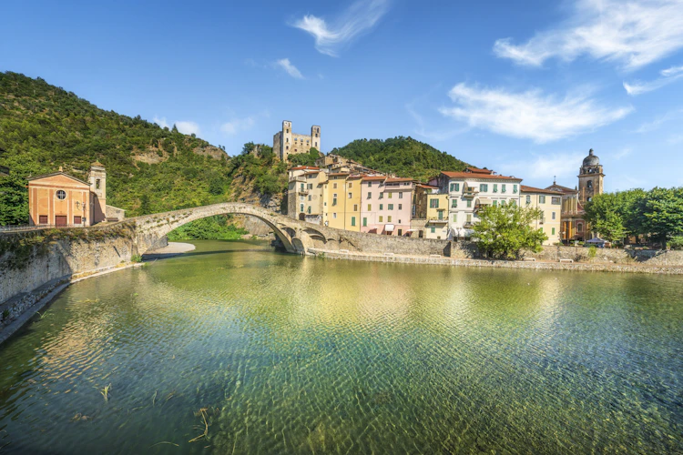 Middelalderlig stenbro over floden Nervia i Dolceacqua, Ligurien, med det historiske Doria-slot på toppen af bakken og farverige italienske huse spejlet i det klare grønne vand