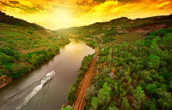 Storslået solnedgang over de terrasserede vinmarker i Douro-dalen, Portugal, med et flodkrydstogtskib der sejler på den snoede flod og jernbanespor langs bredden.