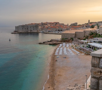 Betagende solnedgang over Dubrovniks historiske bydel og Banje-stranden med den middelalderlige fæstningsby, orange tegltage og det krystalklare Adriaterhav i Kroatien
