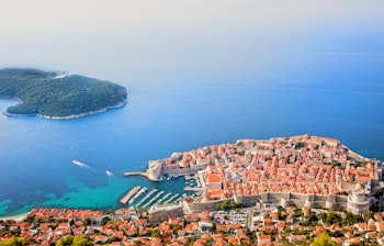 Luftfoto af Dubrovniks historiske gamle bydel med røde tage og den frodige Lokrum-ø omgivet af det azurblå Adriaterhav i Kroatien