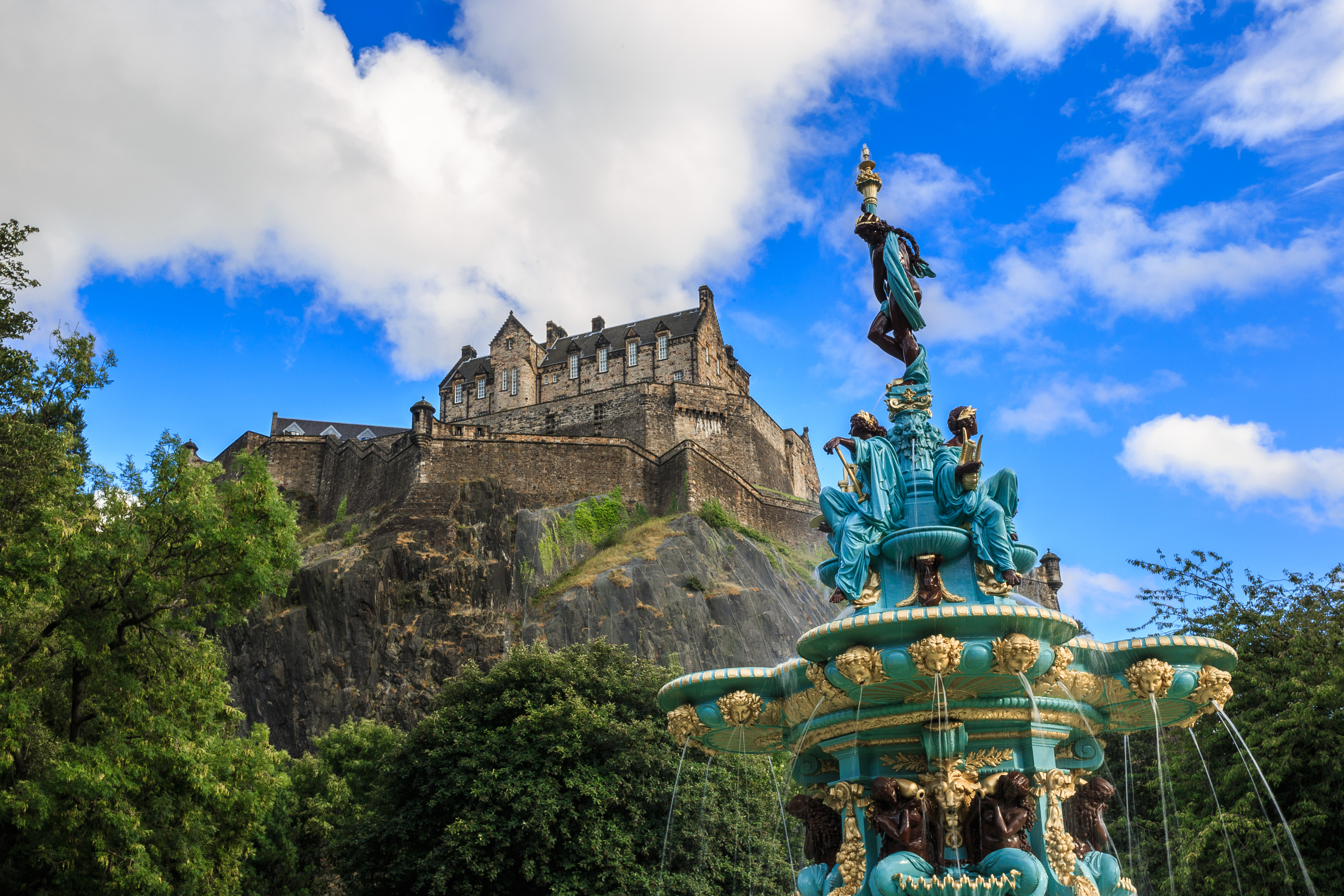 Edinburgh Castle tronende på vulkansk klippe med den smukke Ross-fontæne i forgrunden og blå himmel, Skotland