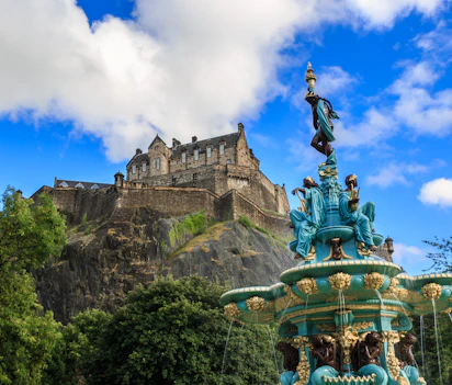 Edinburgh Castle tronende på vulkansk klippe med den smukke Ross-fontæne i forgrunden og blå himmel, Skotland