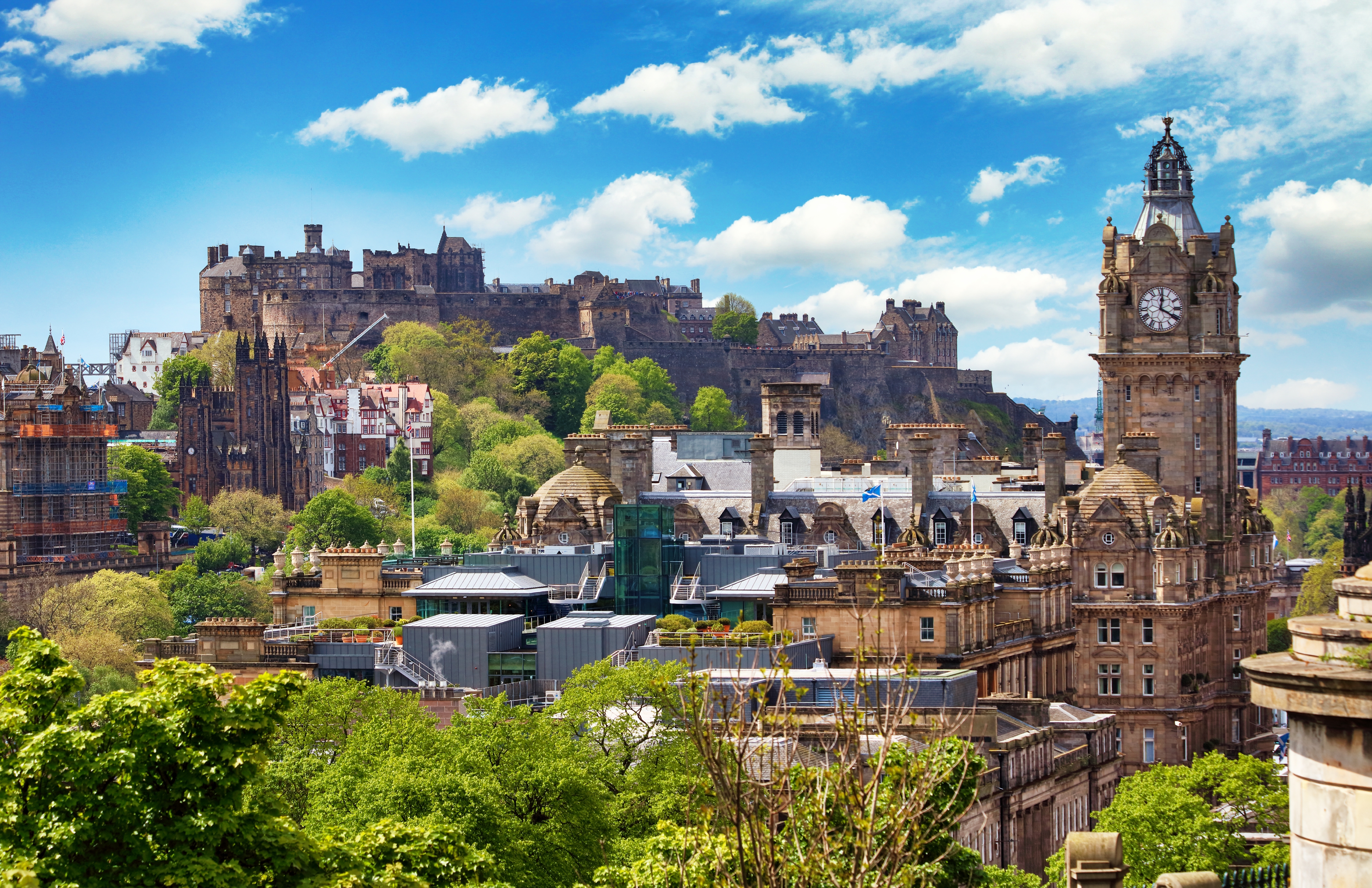 Panoramaudsigt over Edinburgh med Edinburgh Castle på klippetop, Balmoral Hotels klokketårn og historiske sandstensbyggerier under blå sommerhimmel i Skotland