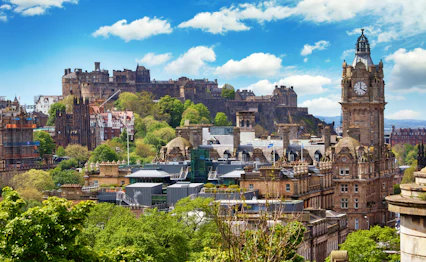 Panoramaudsigt over Edinburgh med Edinburgh Castle på klippetop, Balmoral Hotels klokketårn og historiske sandstensbyggerier under blå sommerhimmel i Skotland