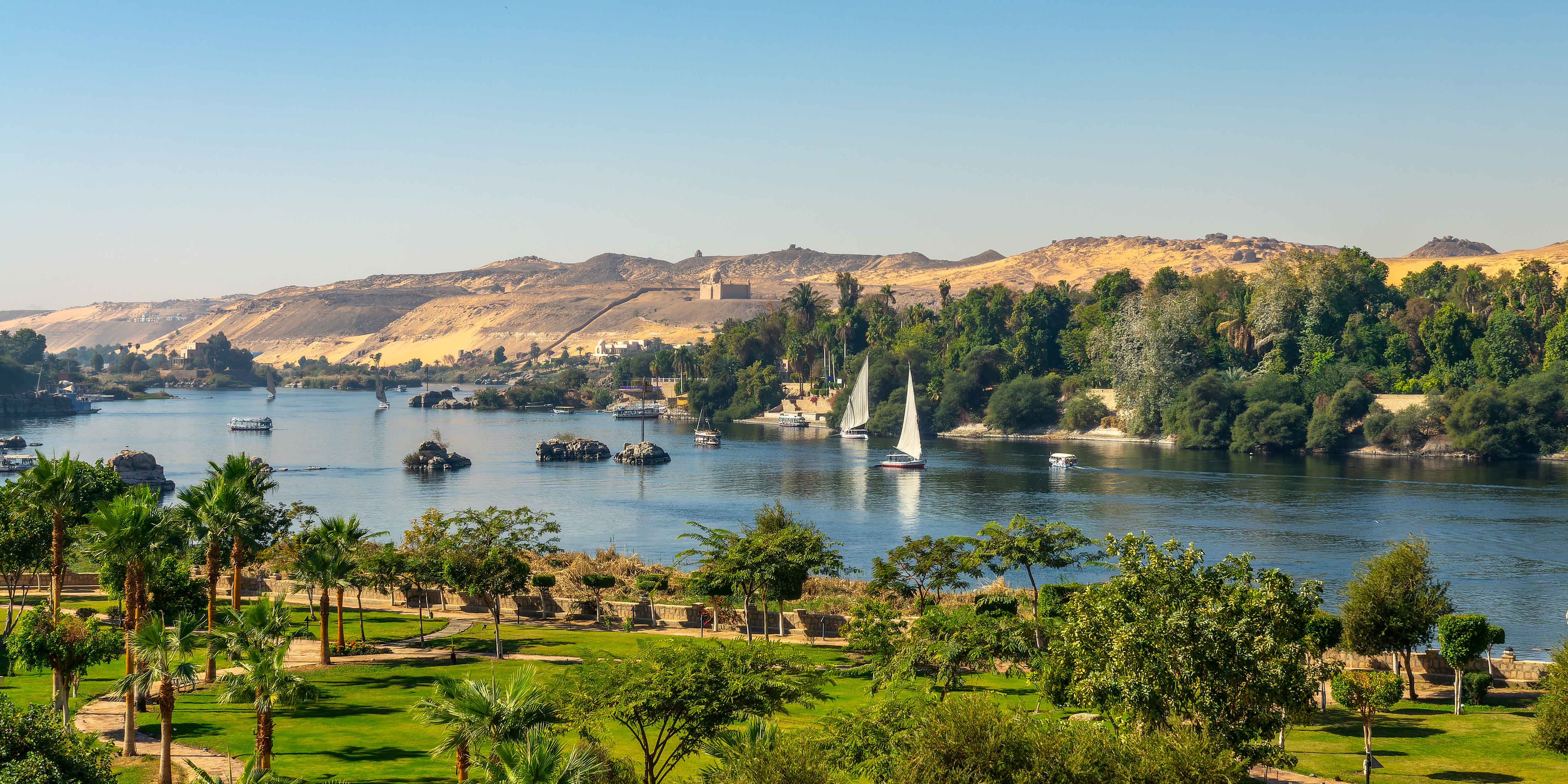 Panoramaudsigt over Nilen i Aswan, Egypten, med traditionelle felucca sejlbåde på det blå vand omgivet af palmetræer og gyldne ørkenklitter