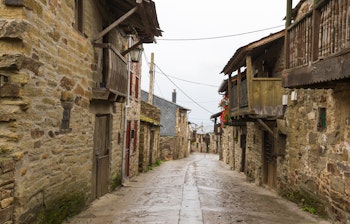 Traditionel stengade i El Acebo på Camino de Santiago pilgrimsruten i León-provinsen, Spanien, med rustikke bygninger og træbalkoner prydet med farverige geranier på en regnfuld dag