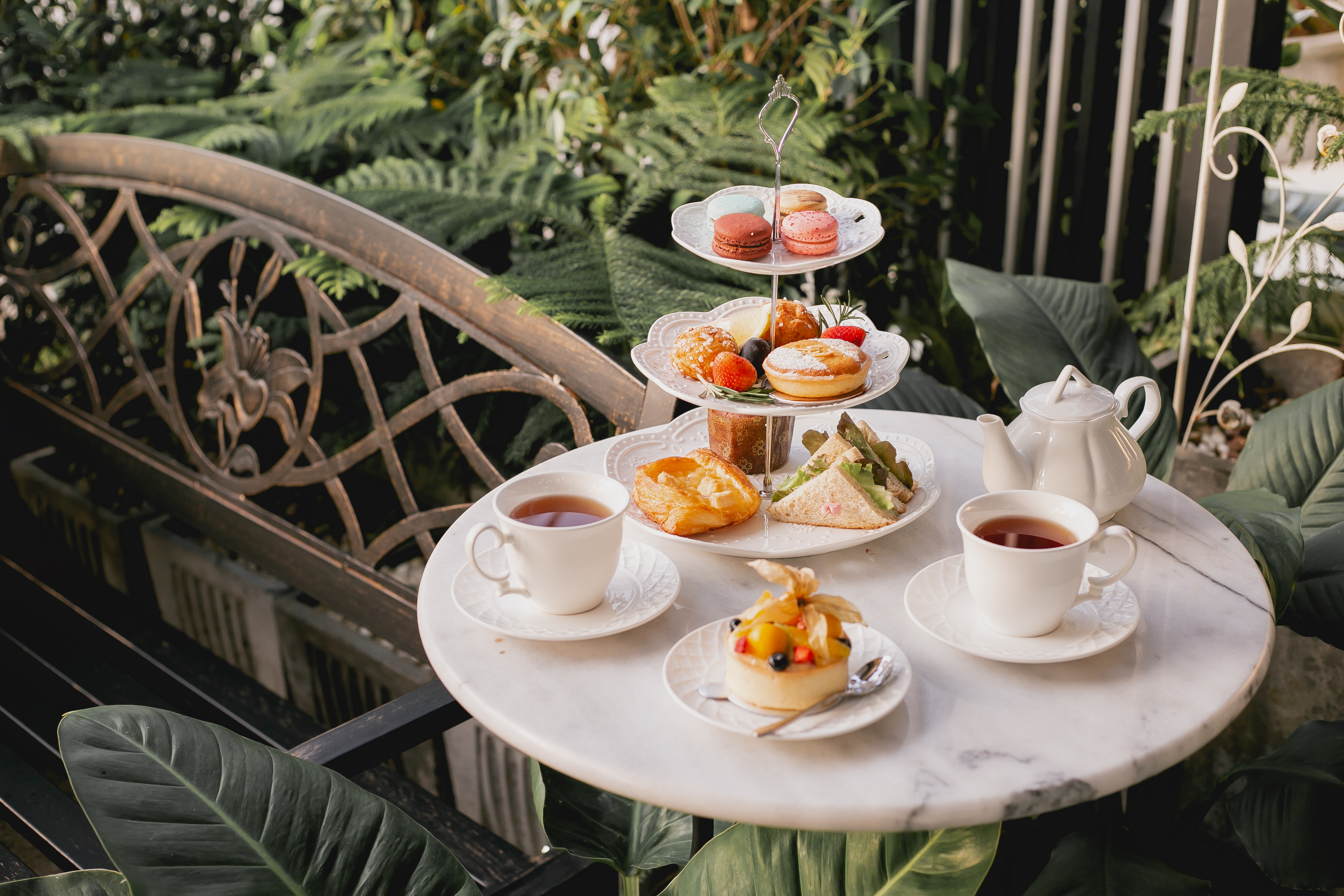 Traditionel engelsk afternoon high tea serveret i en frodig have med tre-etagers kagefad med macarons, kager og sandwiches samt to kopper te