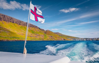 Motorbåd med færøsk flag sejler på Atlanterhavet med Færøernes maleriske kystlinje og bjerge i baggrunden under en klar sommerdag