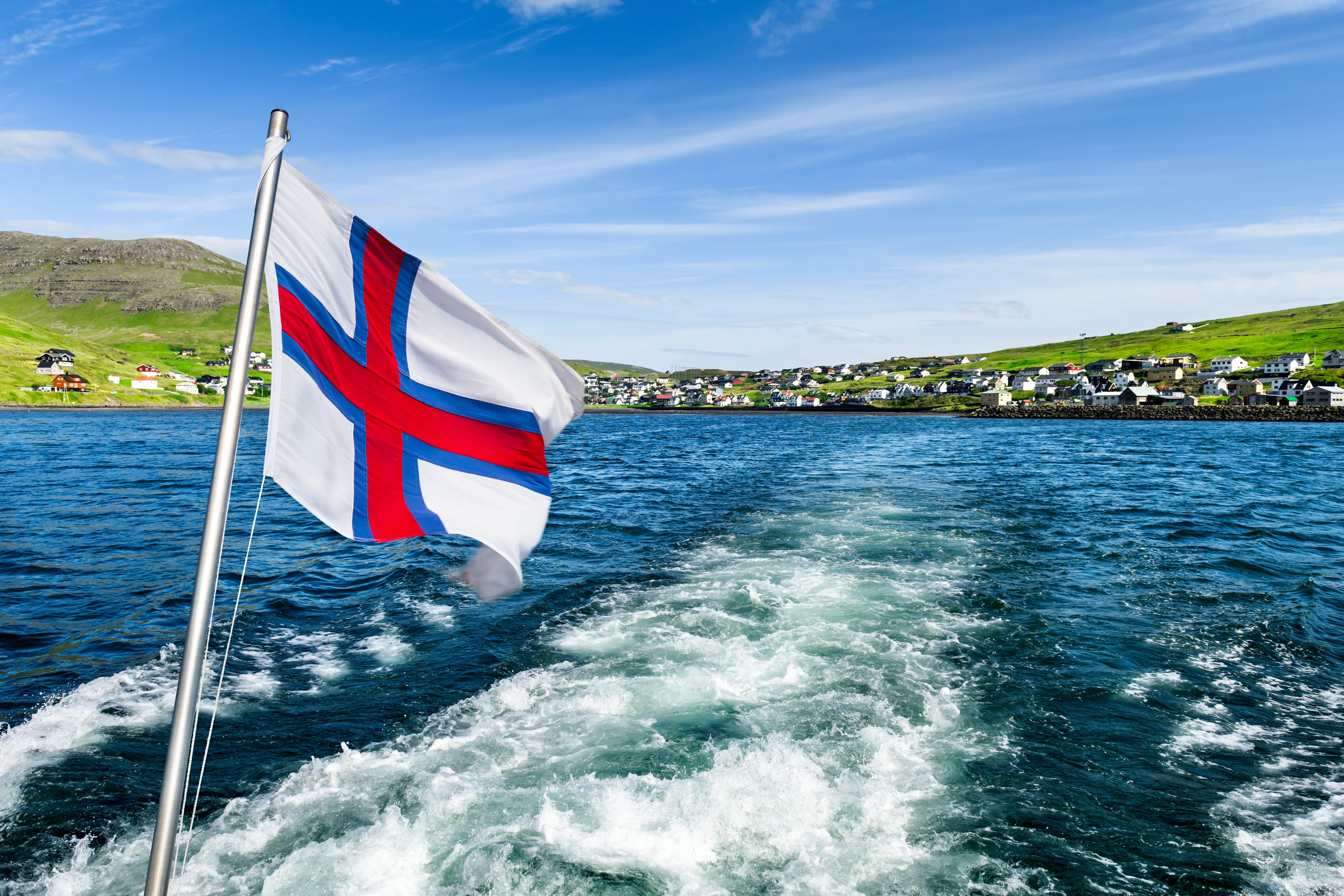 Færøsk nationalflag vajer på en motorbåd på vej fra Sørvágur til Mykines med grønne bakker og kystnær landsby i baggrunden
