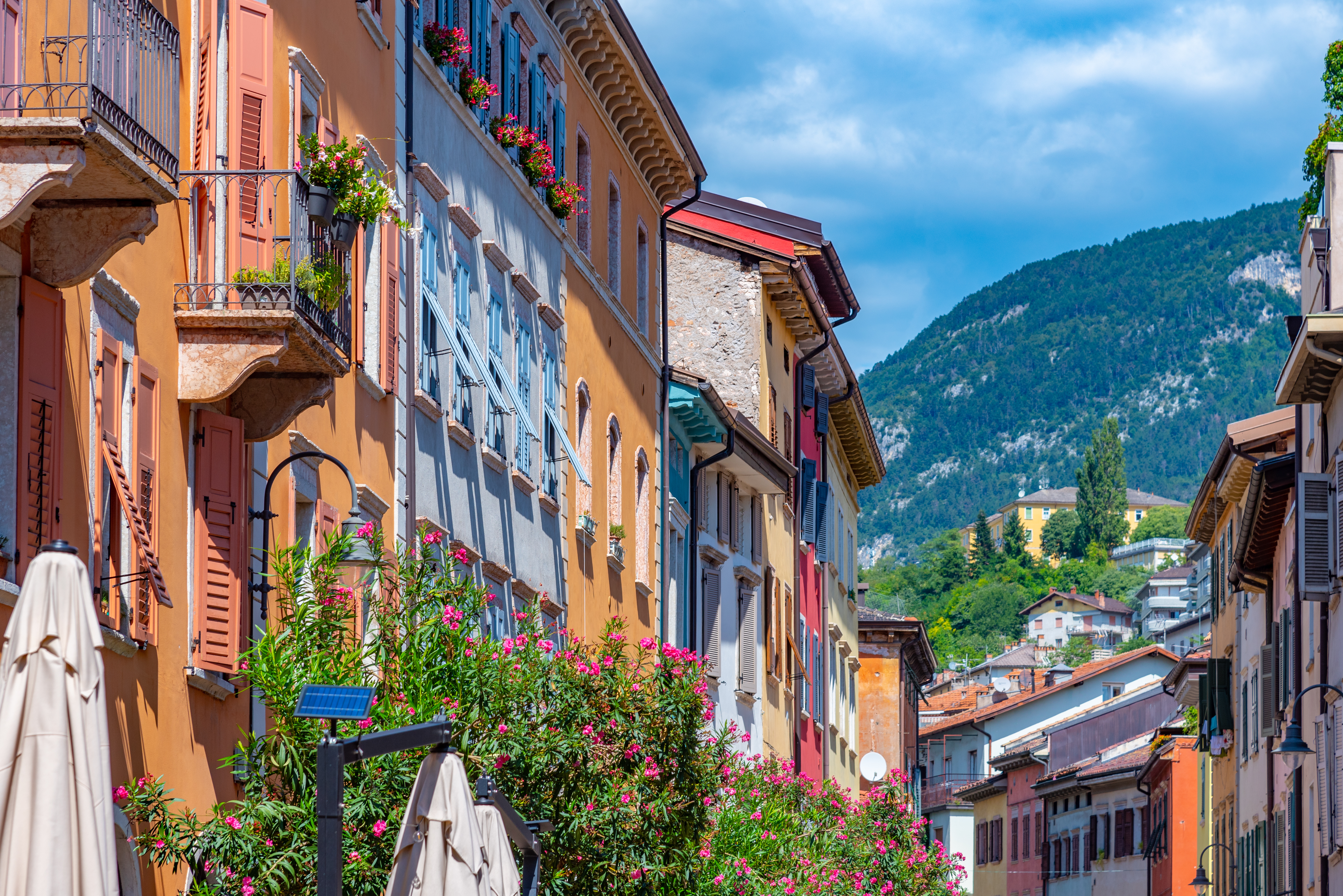 Farverige historiske bygninger langs en smal gade i den gamle bydel i Trento, Italien, med blomsterprydede balkoner og bjerglandskab i baggrunden