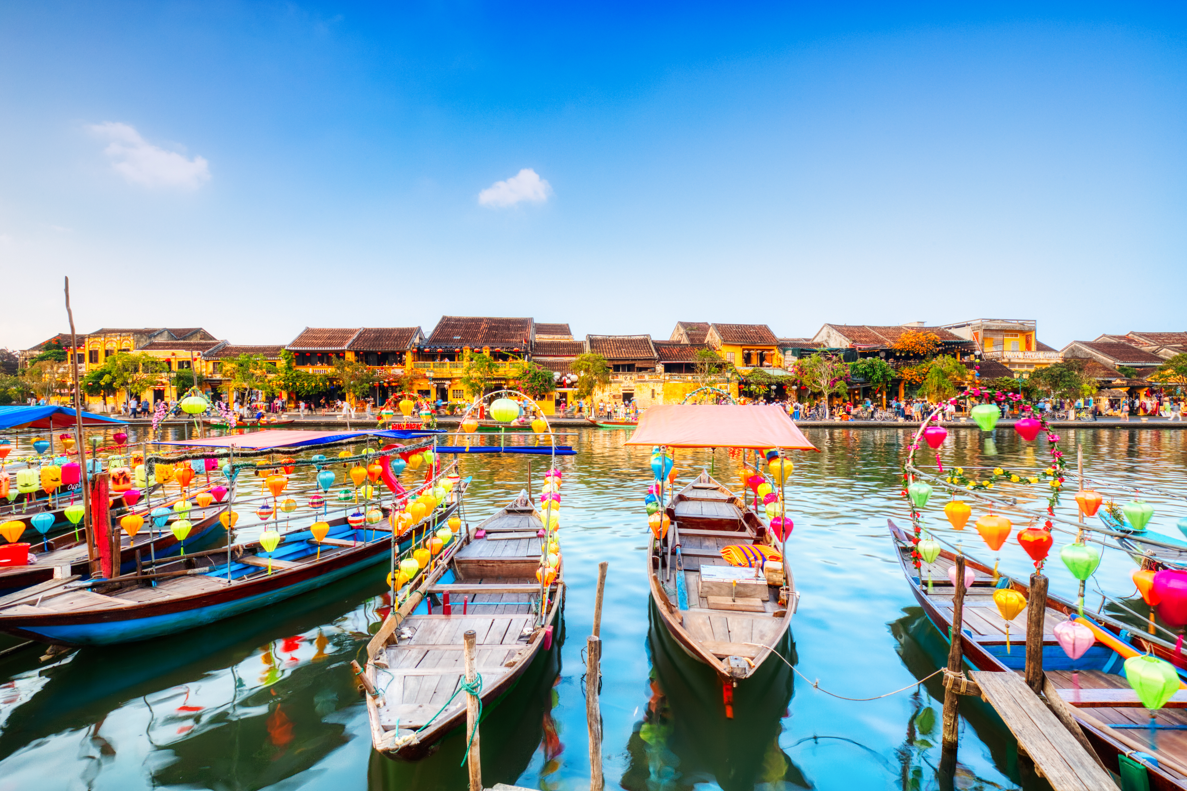 Farverige traditionelle både pyntet med lanterner langs Thu Bon-floden i Hoi Ans gamle bydel i Vietnam med historiske gule bygninger langs vandkanten