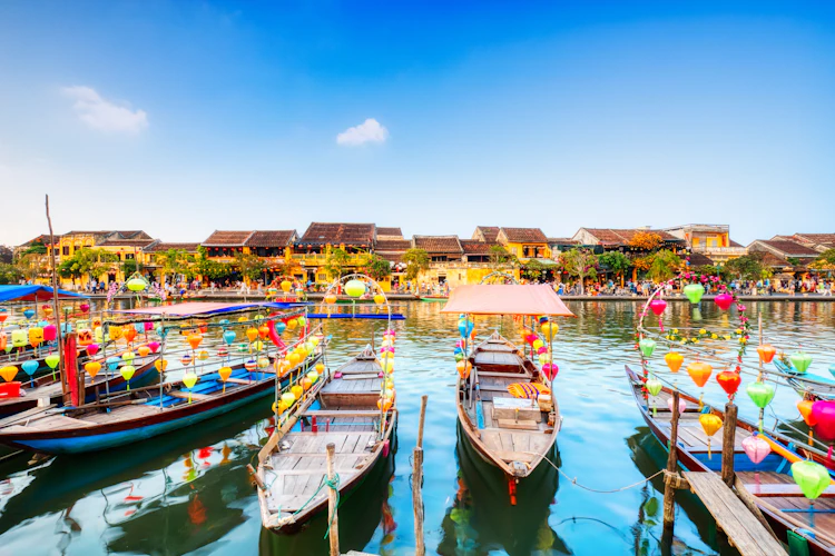Farverige traditionelle både pyntet med lanterner langs Thu Bon-floden i Hoi Ans gamle bydel i Vietnam med historiske gule bygninger langs vandkanten