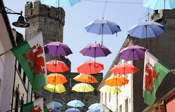 Farverige paraplyer og walisiske flag hænger over Palace Street i Caernarfon, Nordwales, med Caernarfon Slot i baggrunden