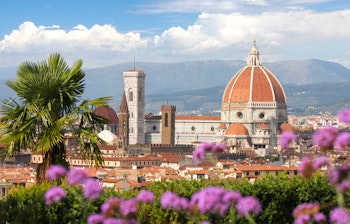Panoramaudsigt over Firenzes domkirke med Brunelleschis kuppel og Giottos klokketårn med lilla blomster i forgrunden og de toscanske bjerge i baggrunden