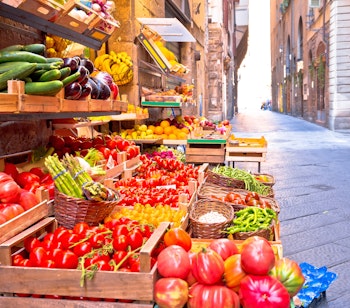 Farverigt frugt- og grøntsagsmarked i en smal historisk gade i Firenze, der viser friske lokale råvarer i trækasser med traditionel toscansk arkitektur i baggrunden