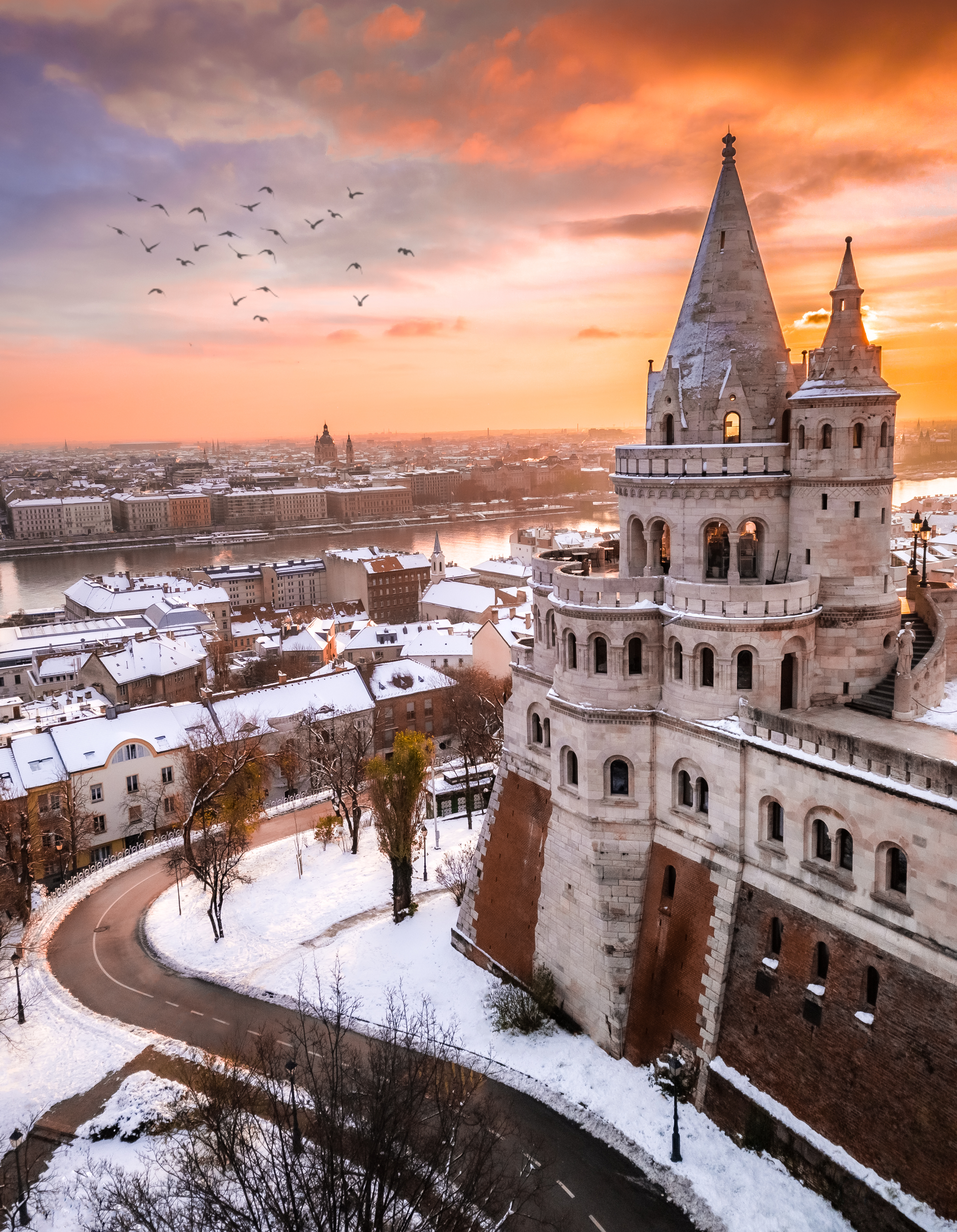 Luftfoto af det sneklædte Fisherman's Bastion i Budapest med Szechenyi-broen og St. Stephens Basilika i baggrunden ved vintersolopgang