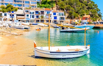 Traditionel fiskerbåd på den gyldne sandstrand i Llafranc med farverige huse langs Costa Bravas kystlinje i Spanien