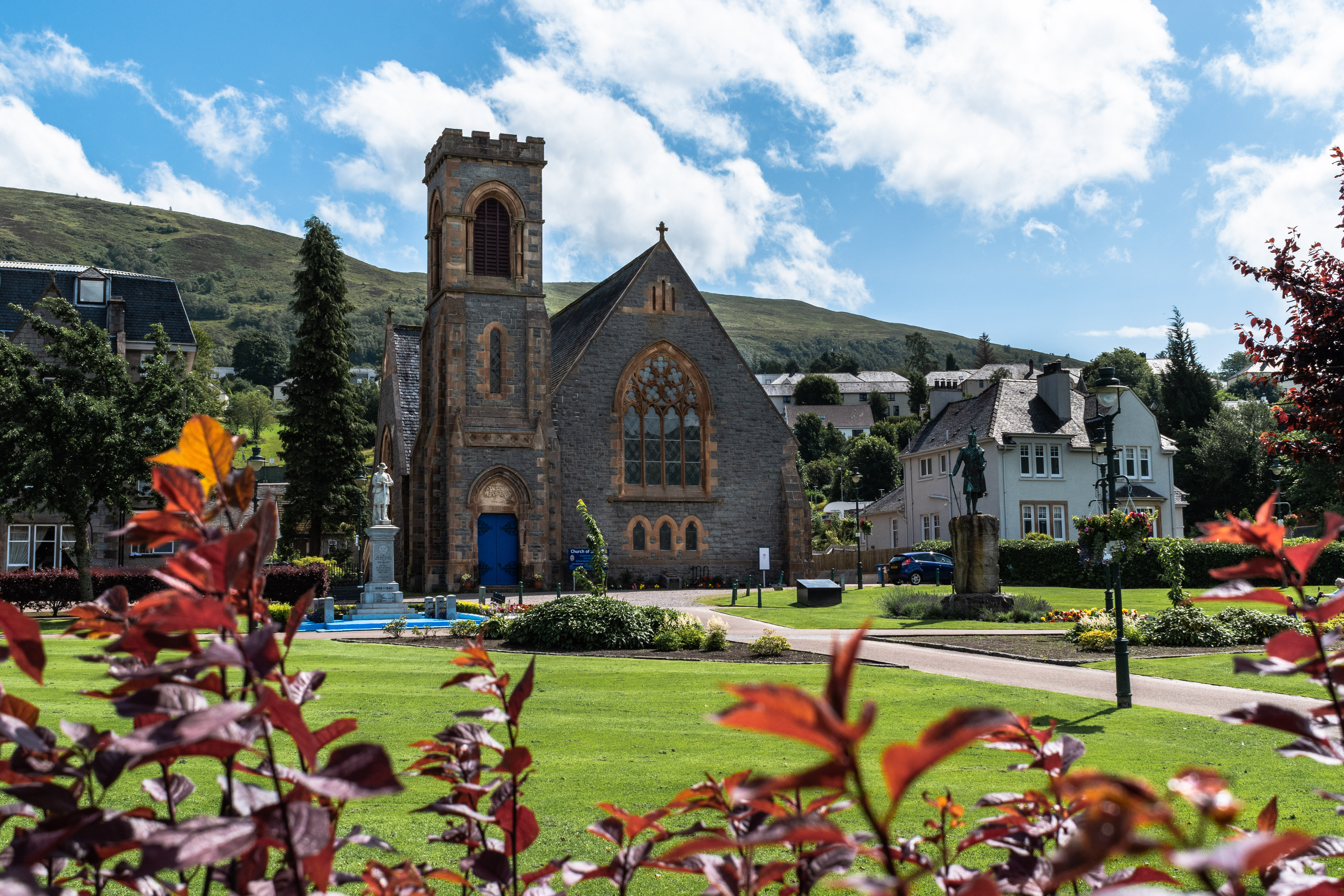 Historisk stenkirke med gotisk arkitektur i Fort William i de skotske højlande, omgivet af grønne haver og røde buske på en solrig sommerdag