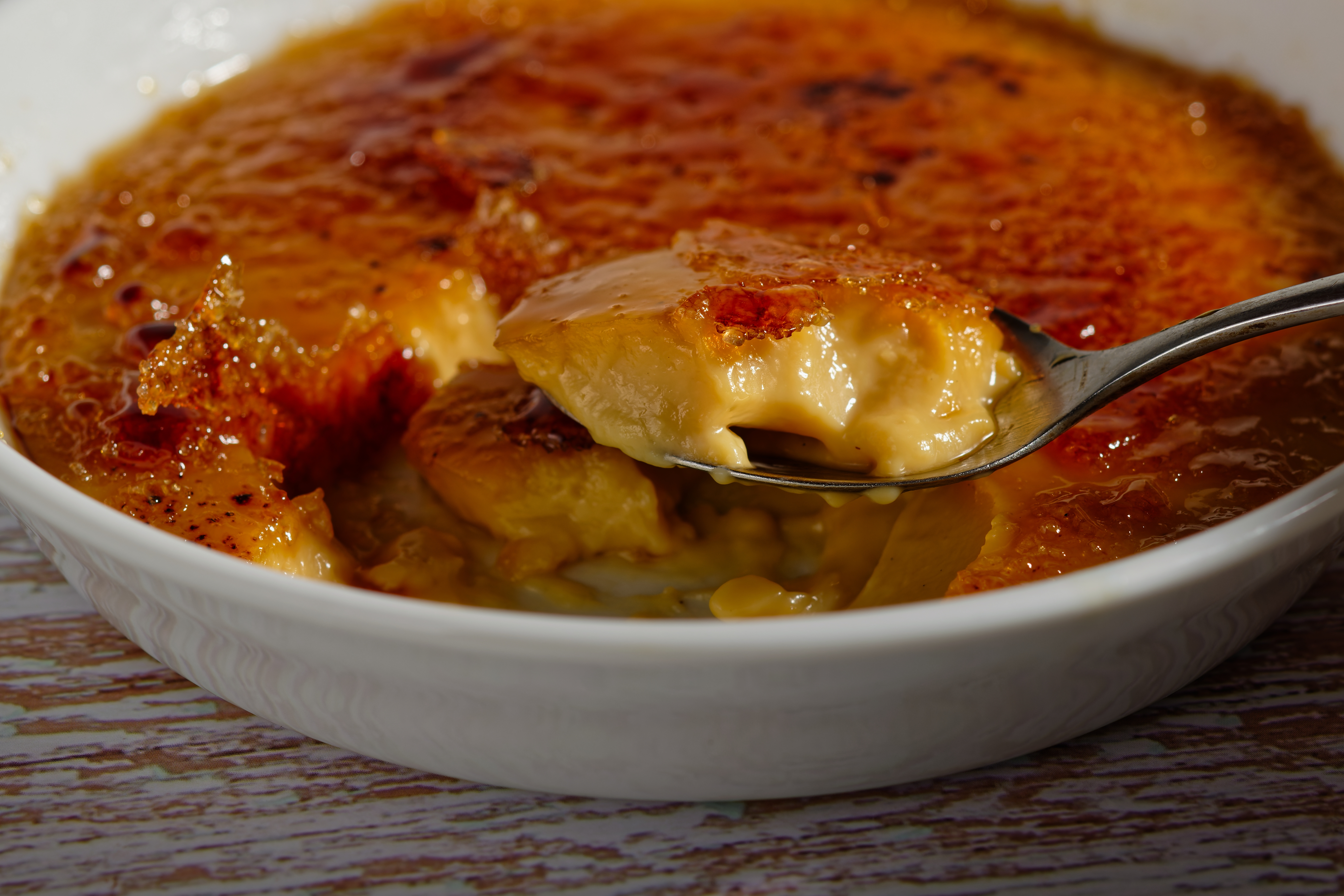 Nært billede af en klassisk fransk crème brûlée med sprød karameliseret sukkeroverside og cremet vaniljecreme, serveret i hvid skål med en ske