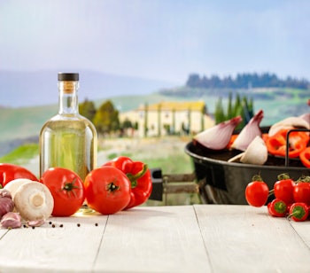 Friske råvarer fra det toscanske køkken med olivenolie, tomater, hvidløg og grøntsager på rustikt træbord med udsigt over de smukke bakker i Toscana