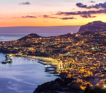 Panoramaudsigt over Funchal by night med oplyste bygninger langs Madeiras kystlinje og bjerge i baggrunden