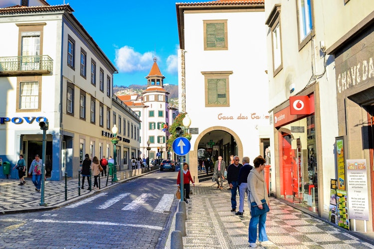 Solrig gade i centrum af Funchal på Madeira med brostensbelagte veje, charmerende portugisisk arkitektur, lokale butikker og turister der udforsker den historiske hovedby under blå himmel