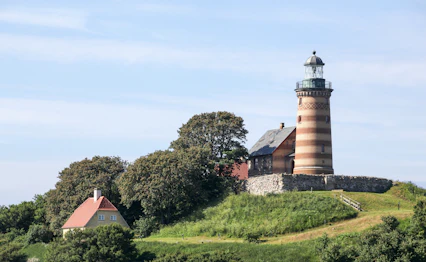 Historisk fyrtårn med røde og hvide striber på grøn bakke omgivet af traditionelle huse og frodige træer