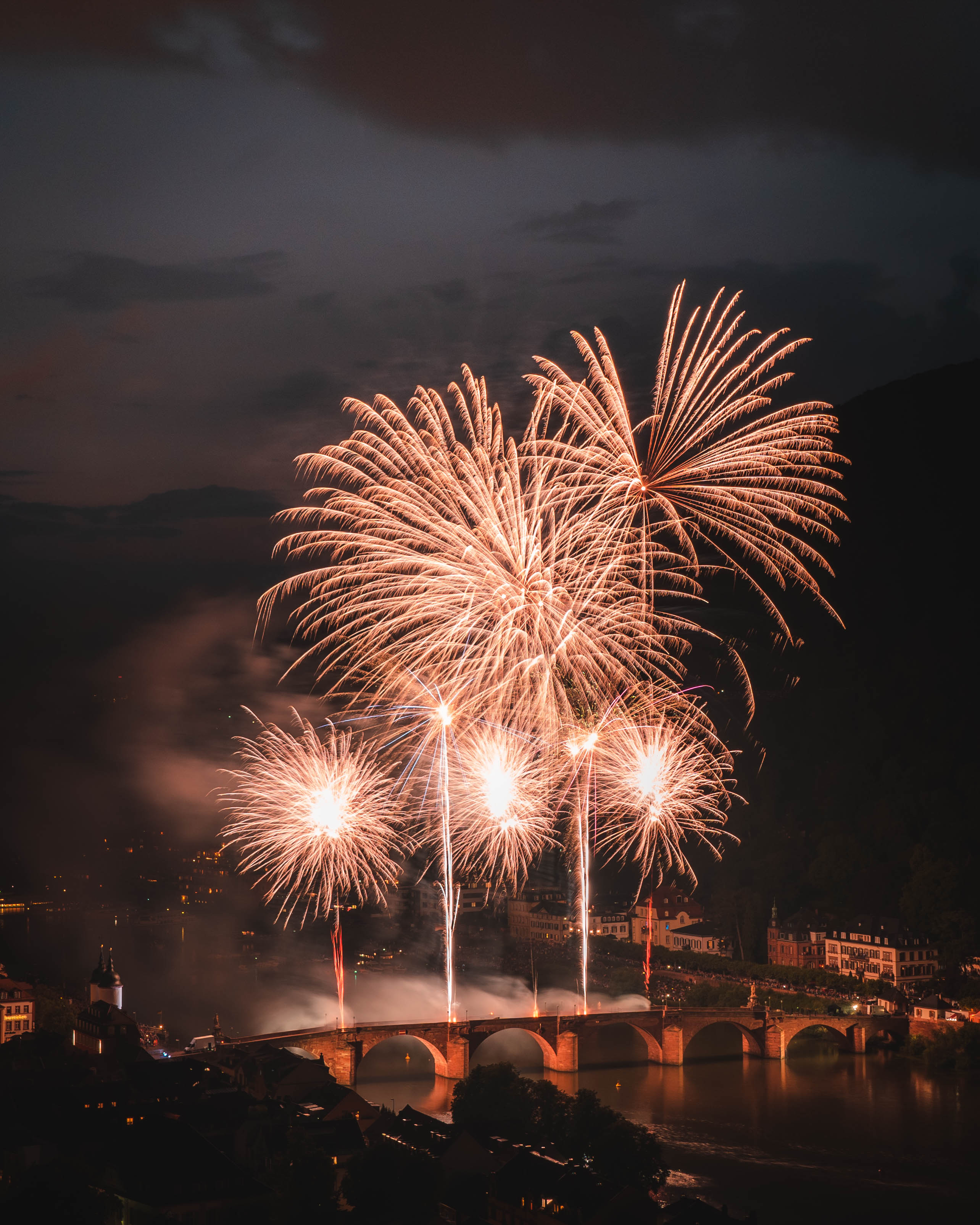 Spektakulært fyrværkeri over historisk stenbro med gyldne eksplosioner der oplyser europæisk bysilhuet om natten