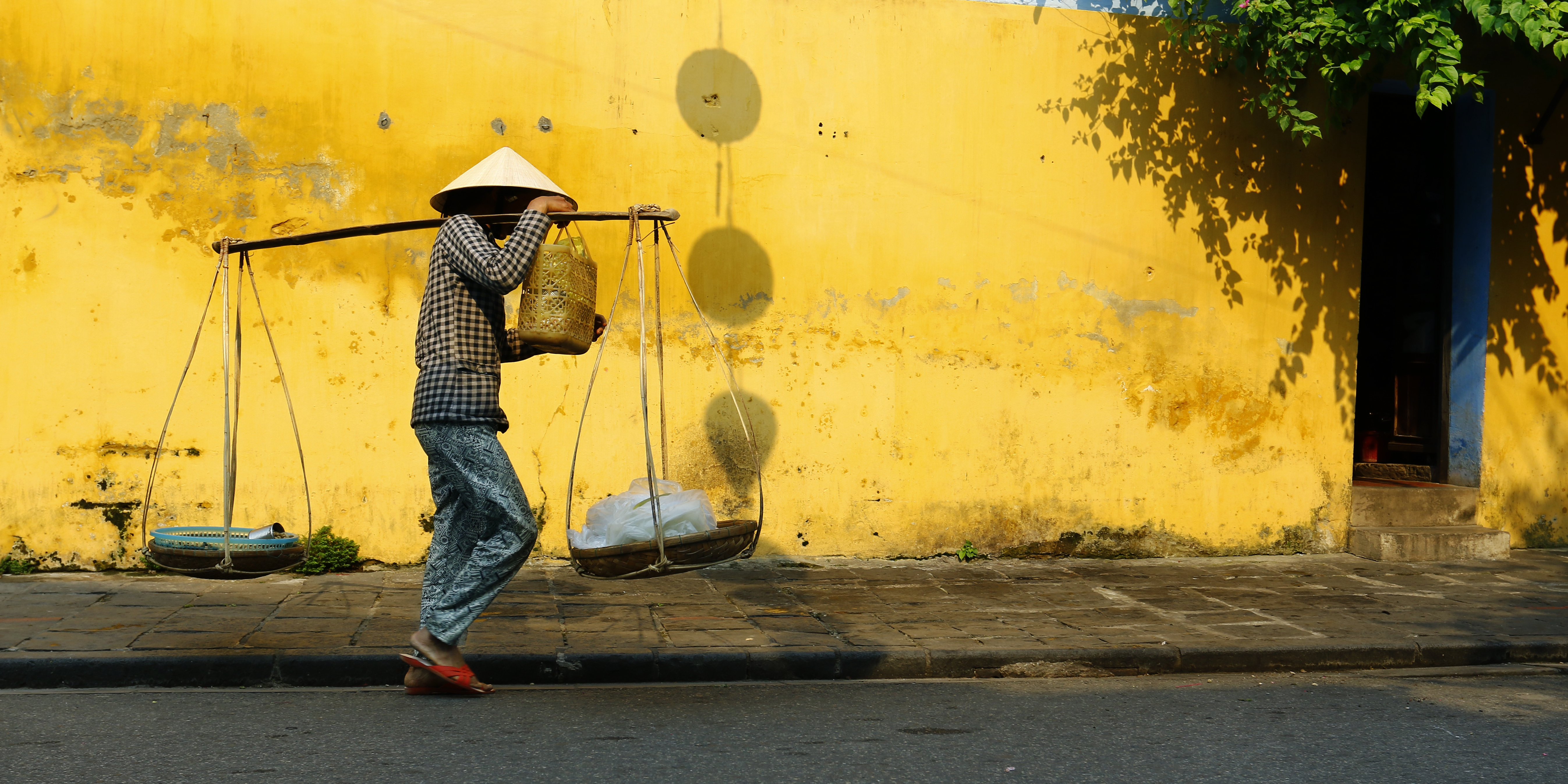 Traditionel vietnamesisk gadesælger med keglehat og bærestang med kurve foran karakteristisk gul mur i Hoi An, Vietnam